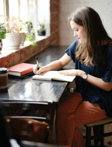 woman sitting in front of black table writing on white book near window